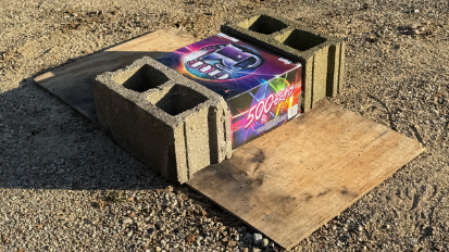 A 500 gram fireworks cake placed on plywood with cement blocks bracing two sides.